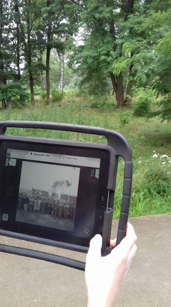 a hand holds the iPad displaying a black and white historical image of Bergen-Belsen contrasting to the empty greenery of the present-day memorial grounds