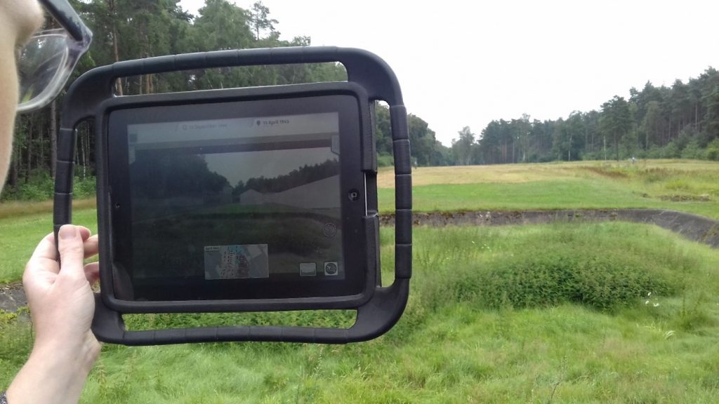 A photograph of a hand holding the iPad with the display of the 3D reconstruction within the present-day memorial site 