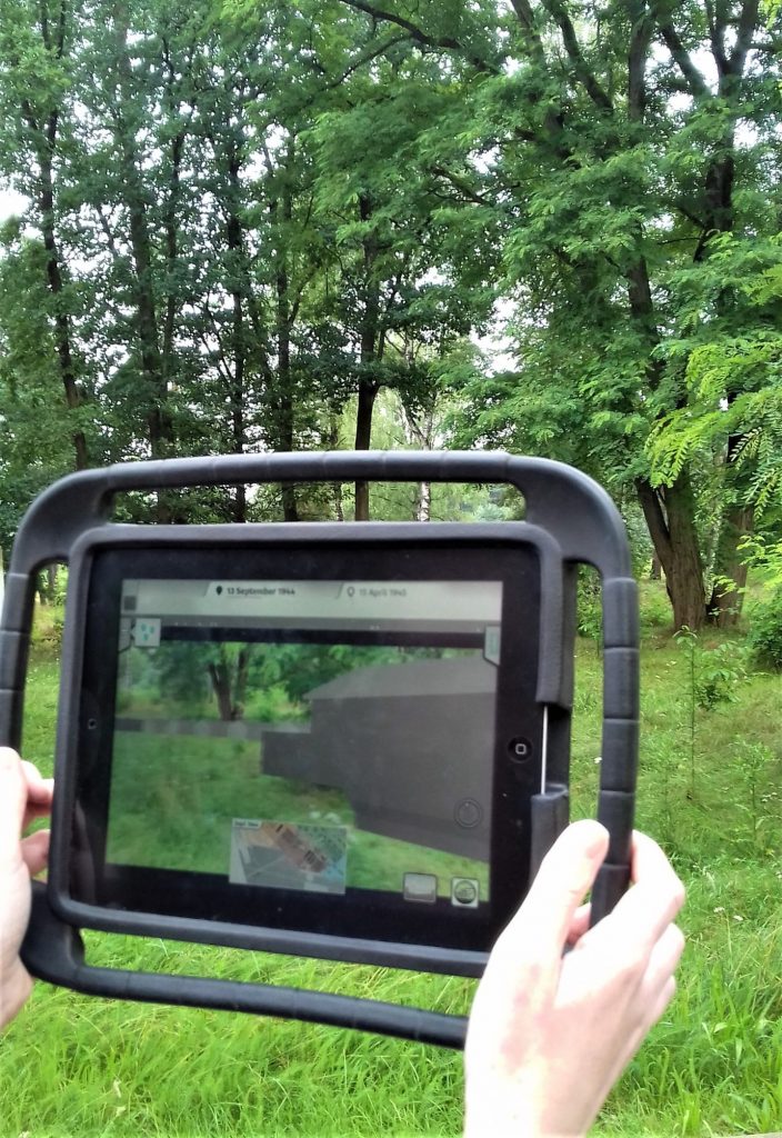 A hand holding the iPad displaying the 3D reconstruction of the barracks overlaid over the present-day memorial site 