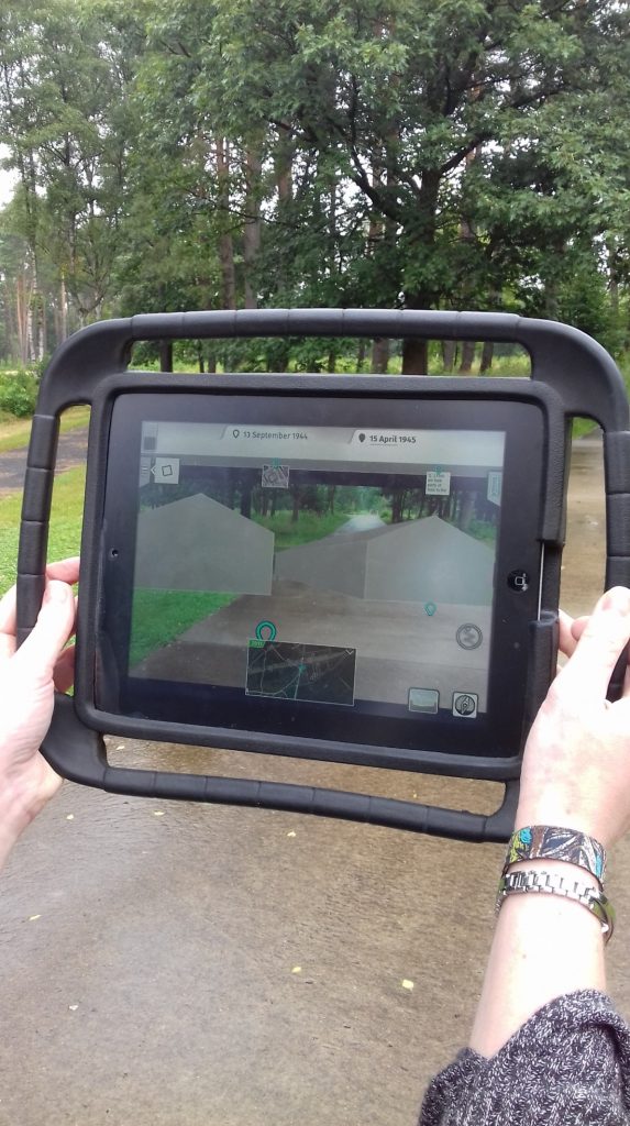 A hand holding the iPad displaying the 3D reconstruction of barracks overlaid over the present-day memorial site pathway