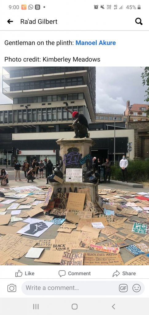 A print screen of a social media post containing a photograph of a person in a protest position sat on top of the plinth formally occupied by Colson surrounded by cardboard signs 