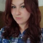 A young woman wit wavy reddish brown hair and a checked blue shirt smirks at an angle.