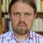 A man with long, tied back blonde hair and a goatee bear wears a checked shirt in front of a blurry bookcase. He stares intently forwards.