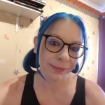 A woman with blue hair in pigtails wearing black glasses tilts her head to the right.