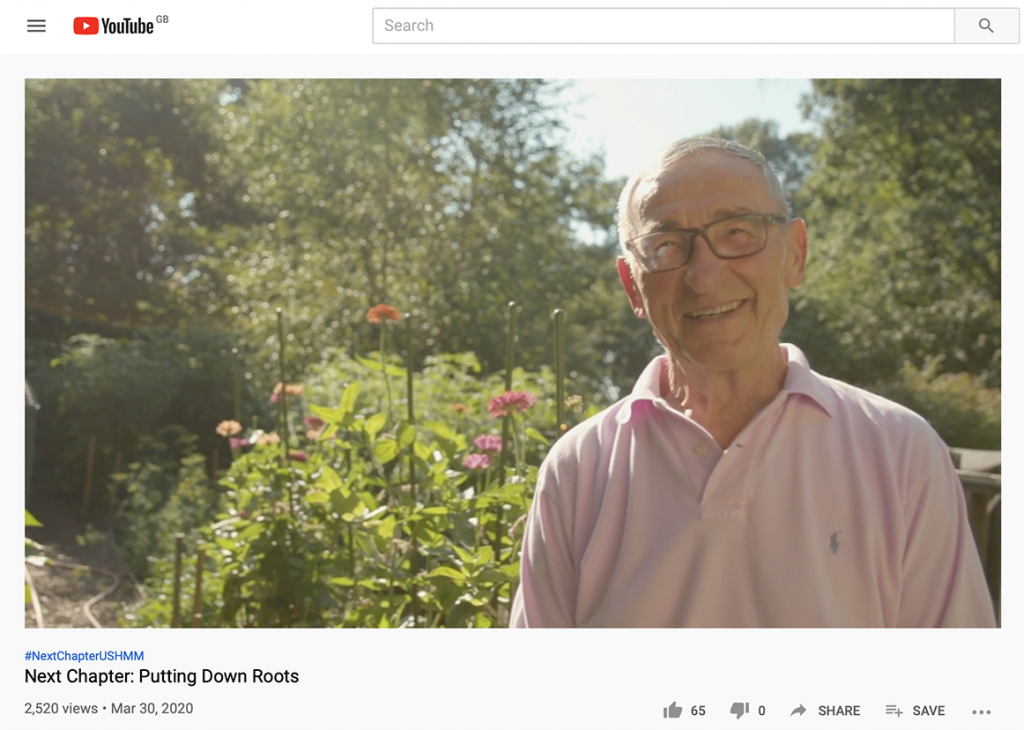 An elderly Holocaust survivor wearing glasses and a pick polo shirt smiles at the camera.