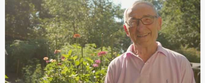 An elderly Holocaust survivor wearing glasses and a pick polo shirt smiles at the camera.