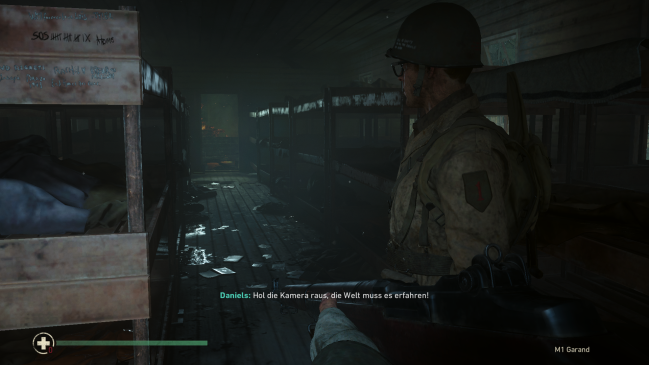 Gameplay screenshot: a soldier in an empty barrack. Text on screen reads: Daniels: Hol die Kamera raus, die Welf muss es erfahren!