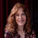 A woman with wavy auburn hair, glasses and a smart dress smiles.