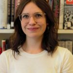 A young woman with mid-length dark-brown hair and glasses. She wears a white t-shirt and smiles. She is stood in front of a bookcase.