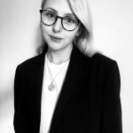 Young woman with blonde hair and glasses. She wears a smart casual suit and looks forward.