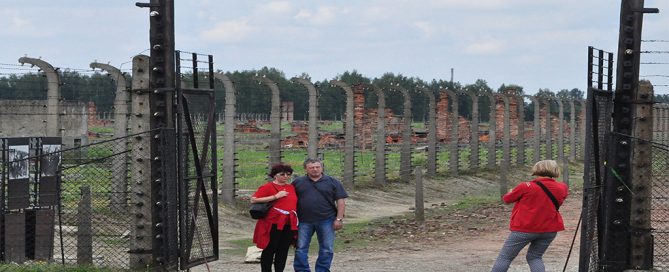 The image shows a scene at a concentration camp site, with several visitors standing near the entrance, framed by a pair of tall, barbed-wire-lined gates. In the background, remnants of the camp's structure, including concrete posts and barbed-wire fences, stretch into the distance, creating a somber and reflective atmosphere. The visitors are engaged in taking photos, likely capturing memories of this historically significant and solemn place. The image serves as a reminder of the importance of preserving history and fostering remembrance through education and visitation.