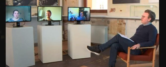 A man looks at three screens on stands - each has a presenter on.