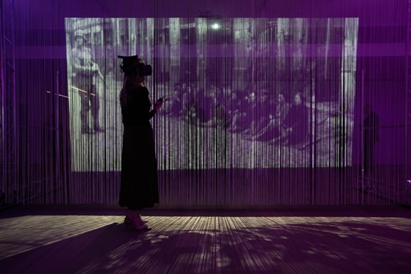 A projection of a historical photograph stands behind a woman with a VR headset on.