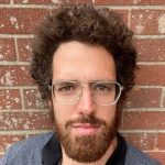 A man with bushy hair, a beard and glasses looks directly at the camera.