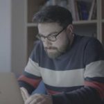 A man with fluffy hair, a beard and glasses stares intently at a laptop.