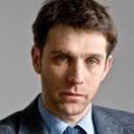 A man with short fluffy hair wearing a blue and grey suit looks downwards with a serious gaze.