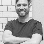 A man with scruffy hair and a huge smile wears a t-shirt and has his arms crossed.