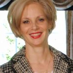 A woman with short blonde hair wears a smart jacket and smiles.