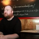 A cheery man in a black t-shirt, with very short hair and a bear sits in front of a black board.