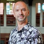 A bald man with stubble wears a flowery shirt and looks at the camera.