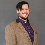 A man smiles enthusiastically for the camera. His short hair slightly drapes over his face. He wears a brown jacket and checked blue and purple shirt.