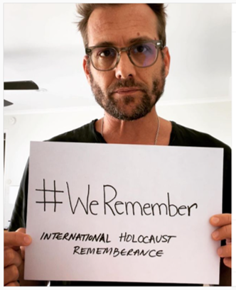 A man with dark hair, beard and glasses holds a sign reading: hashtag we remember, international holocaust remembrance.