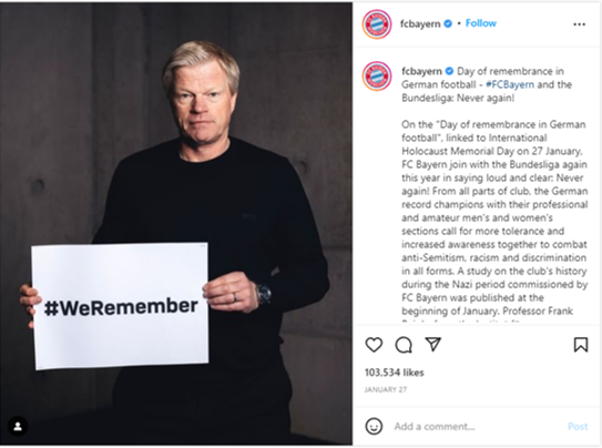 A well-built man with short blonde hair wearing all black, holds a sign saying hashtag we remember.