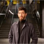 A young man with a beard wears a black overcoat and black jumper. He stands in front of a large staircase. 