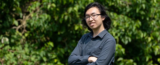 A young man, arms crossed and wearing glasses and a shirt smiles against a leafy backdrop.