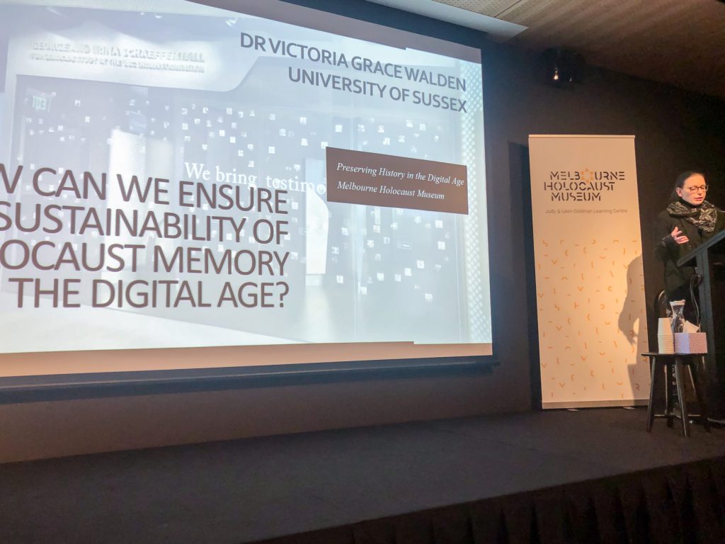 Woman with blue hair stands at a lectern in front of a presentation that reads 'How Can We Ensure Sustainability of Holocaust Memory in the Digital Age?'