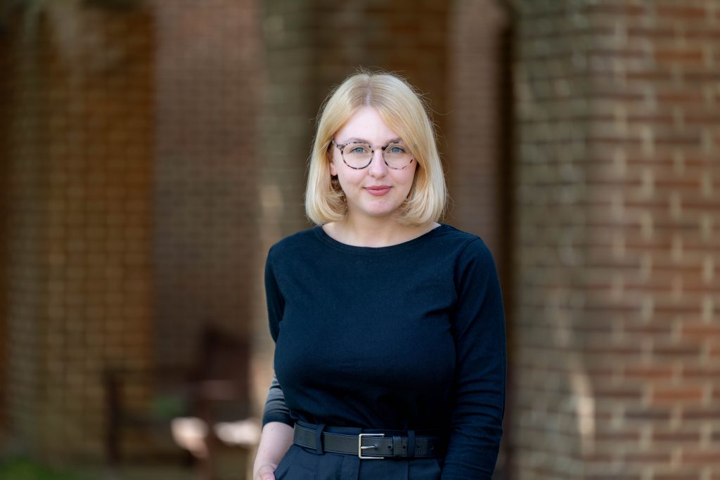 A young blonde woman with glasses, dressed all in black stands against a brick background.