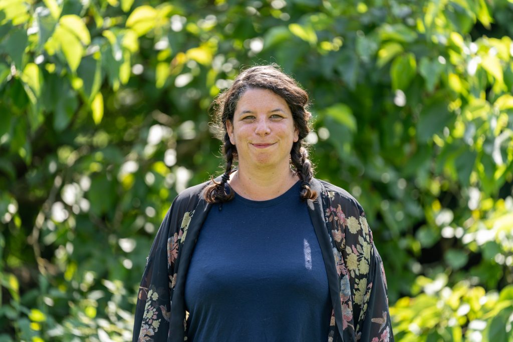 A woman with pigtails in her hair smiles at the camera, against a leafy backdrop. 