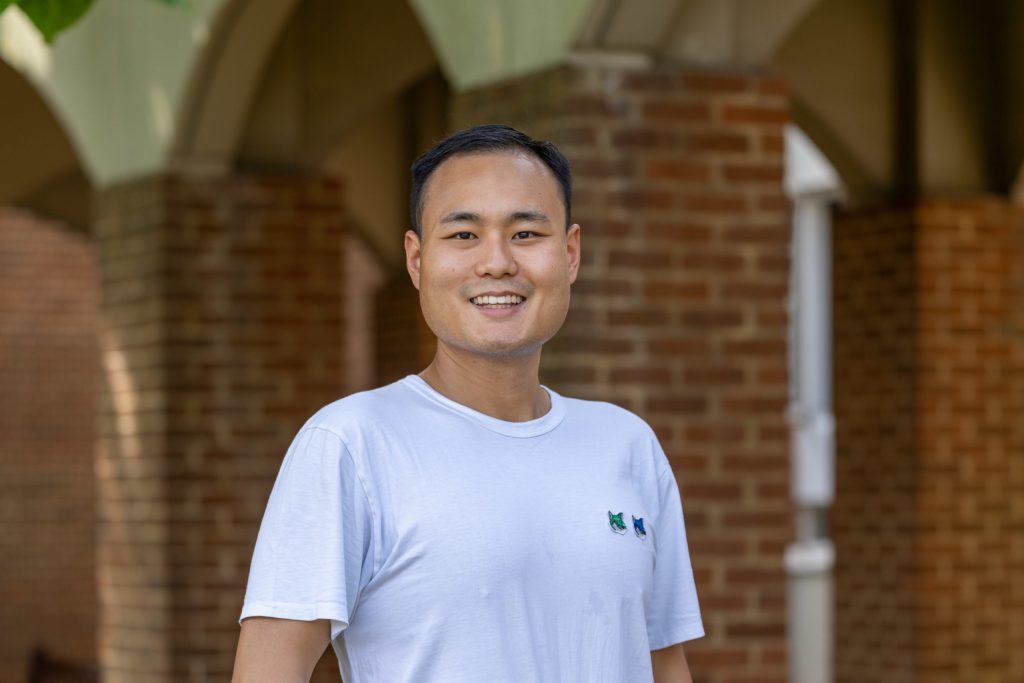 A young man stands proudly in a white t-shirt, smiling, against a brick background.