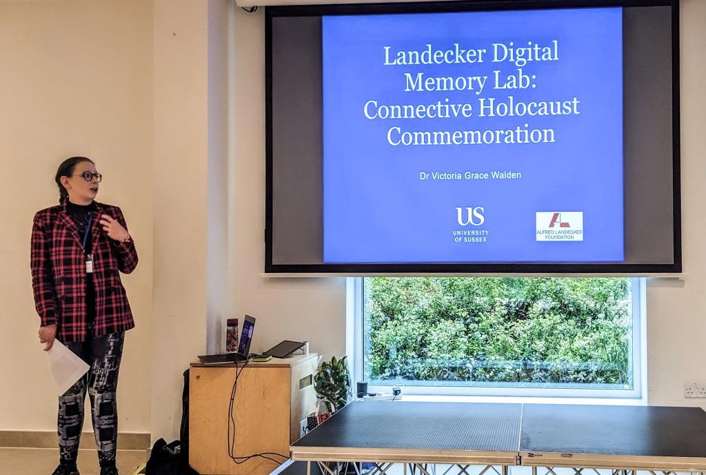 A woman with blue hair speaks to the side of a presentation which says 'Landecker Digital Memory Lab: Connective Holocaust Commemoration'.