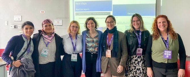 seven women in casual dress pose - smiling - in front of a PowerPoint presentation.