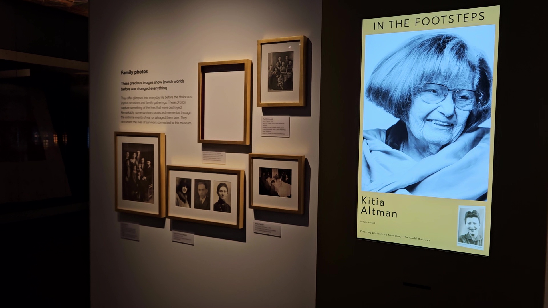 A digital screen that says 'In The Footsteps' and displays a black and white photograph of an elderly woman - a Holocaust survivor - stands next to a traditional museum display of framed photographs