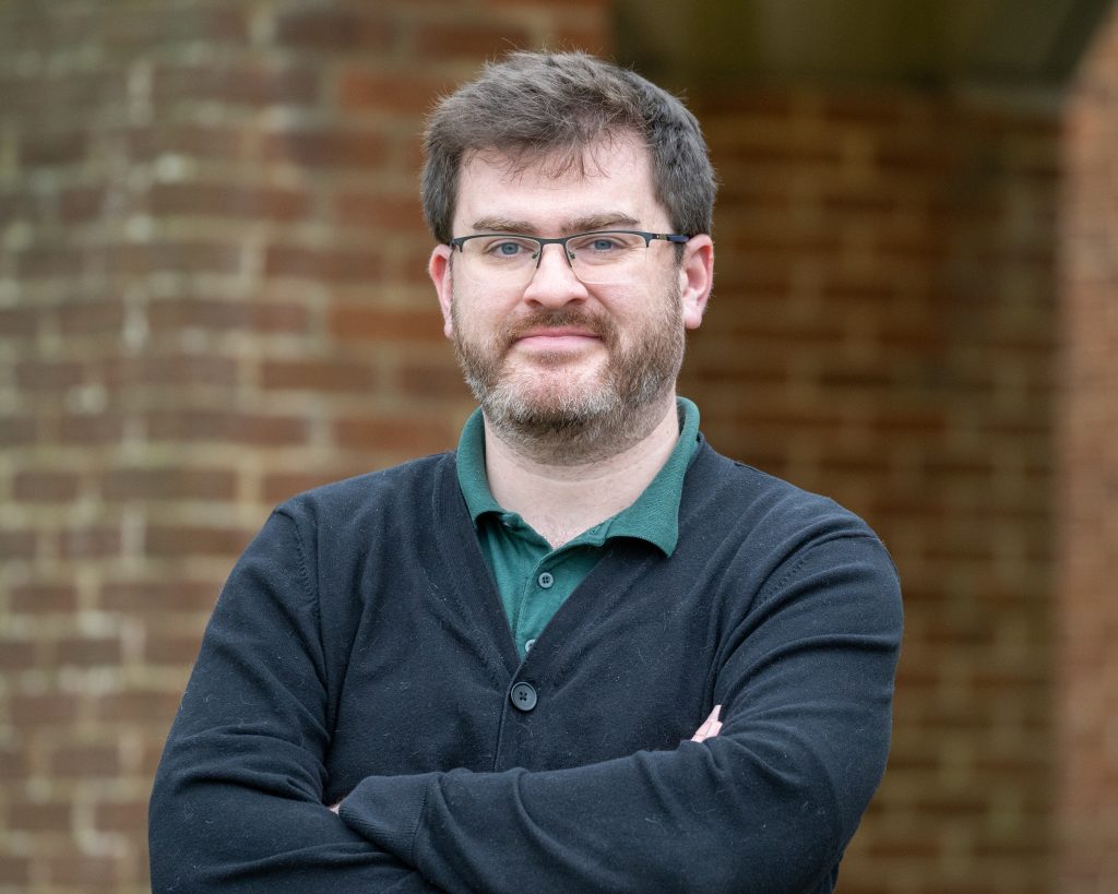 The photo shows an individual with short dark hair, glasses, and a beard. They are wearing a dark cardigan over a green collared shirt and have a confident, approachable expression. The background features a brick wall, adding a warm, rustic feel to the setting. Their arms are crossed, giving a relaxed yet professional posture.