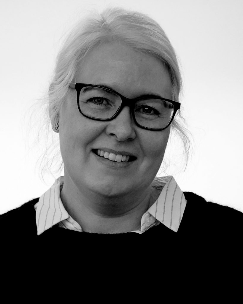 A person with light hair, wearing glasses, smiles warmly in this black-and-white portrait. They are dressed in a collared shirt under a sweater, and the plain background keeps attention on their friendly expression, giving a professional yet approachable impression.