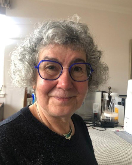 A person with curly gray hair and distinctive blue glasses smiles warmly in this indoor portrait. They are dressed in a dark top, and the background shows a casual setting with various household items, adding a friendly and approachable feel to the image.