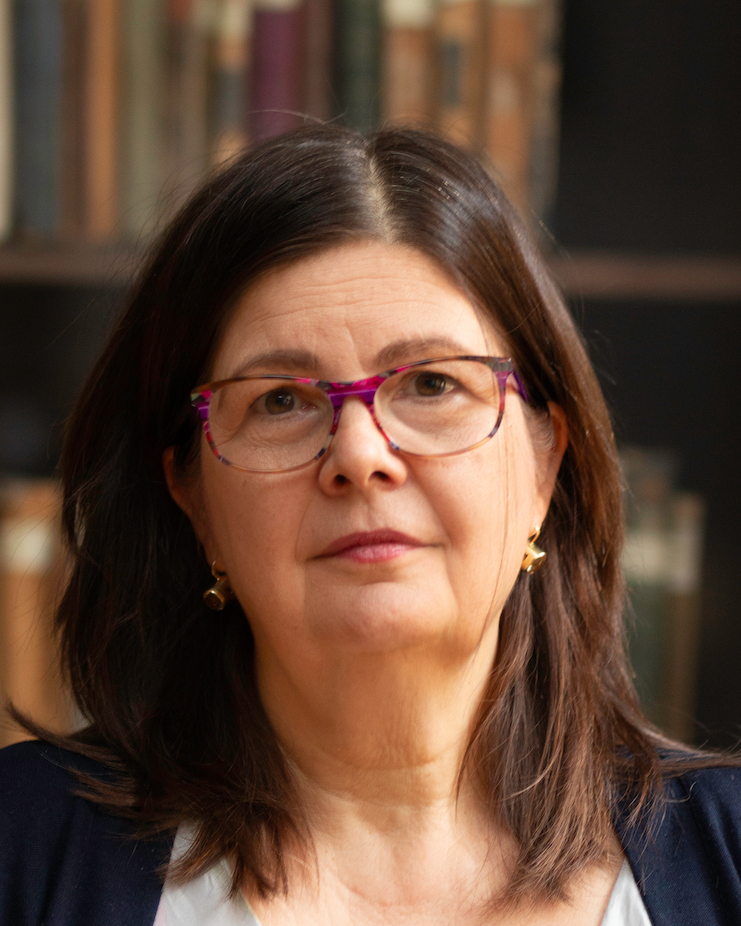 A person with shoulder-length dark hair, wearing glasses with a distinctive purple frame. They have a calm and composed expression, and the background includes shelves of books, adding an intellectual and academic ambiance to the image. They are wearing earrings and appear dressed in professional attire, with a thoughtful, focused demeanour.