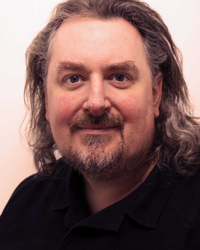 A man with long wavy hair and a beard is shown in a close-up portrait. He has a calm expression with gentle eyes and a slight smile, wearing a dark shirt. The background is simple, keeping the focus on his face.