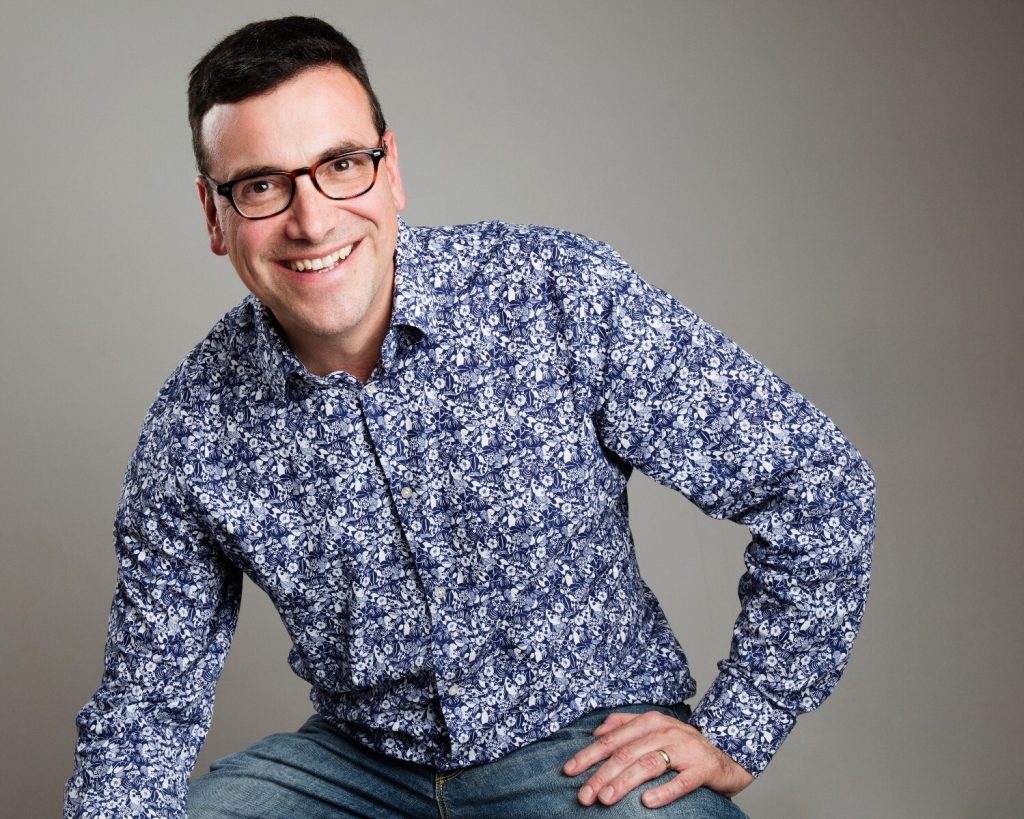 A person with short dark hair and glasses smiles warmly in this portrait. They are wearing a patterned blue shirt and jeans, leaning slightly forward with a relaxed and friendly demeanor. The plain background emphasizes their approachable and confident expression.