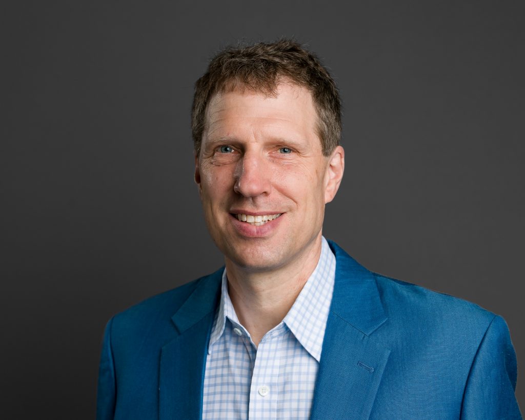 This image shows a portrait of a person in a blue suit jacket and checkered shirt, posing against a dark gray background. They are smiling and looking directly at the camera.