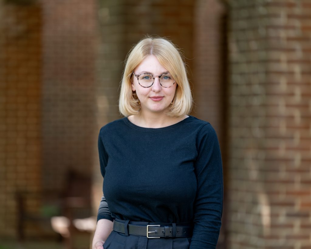 A person with short blonde hair, wearing glasses and a black outfit, stands outside in front of a brick building. She has a calm, composed expression, and the background shows soft, natural light with blurred brick arches.