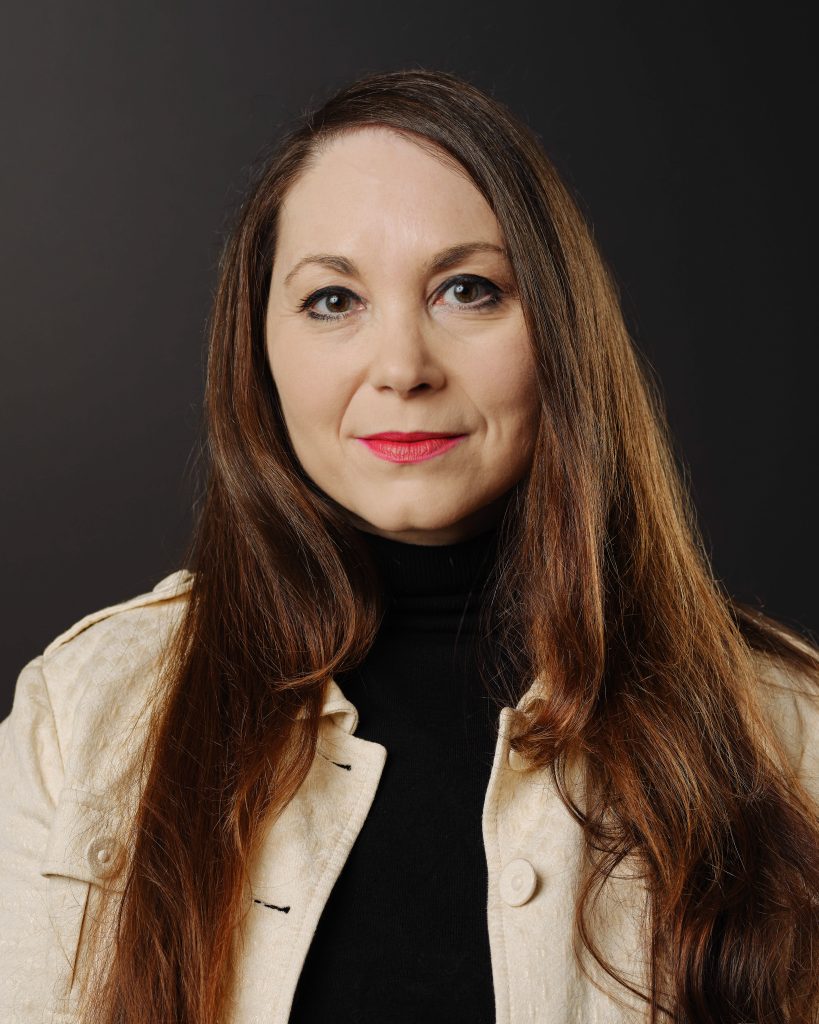 A person with long brown hair and a confident expression is shown in a portrait against a dark background. They are dressed in a light-colored jacket over a black turtleneck, with a touch of bold red lipstick adding contrast. The simple background highlights their composed and professional demeanour.