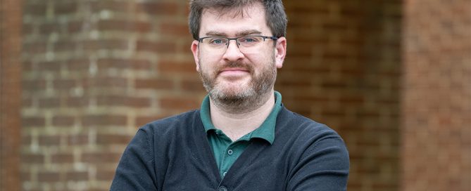 A man with fluffy brown hair, beard and glasses stans with his arms crossed wearing casual clothes in front of a blurry brick wall.