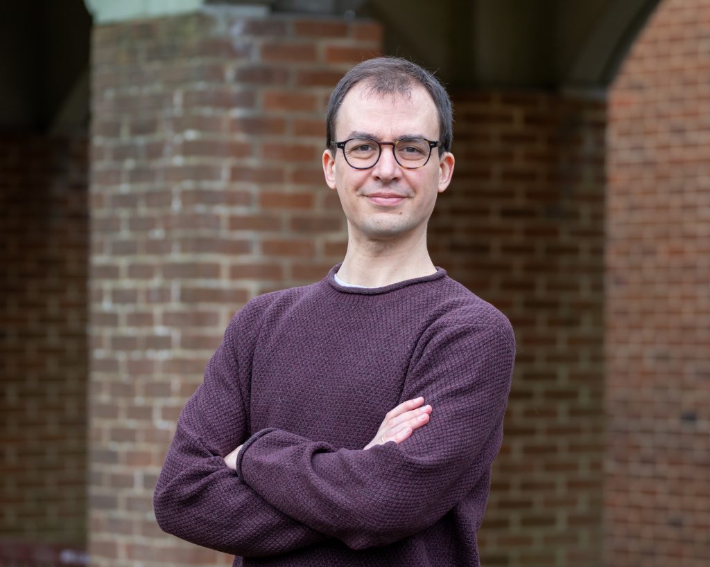 This image shows a man with short hair and glasses, wearing a maroon sweater and standing with arms crossed. He has a composed expression, and the background includes a brick wall and an archway, providing an academic or formal outdoor setting.