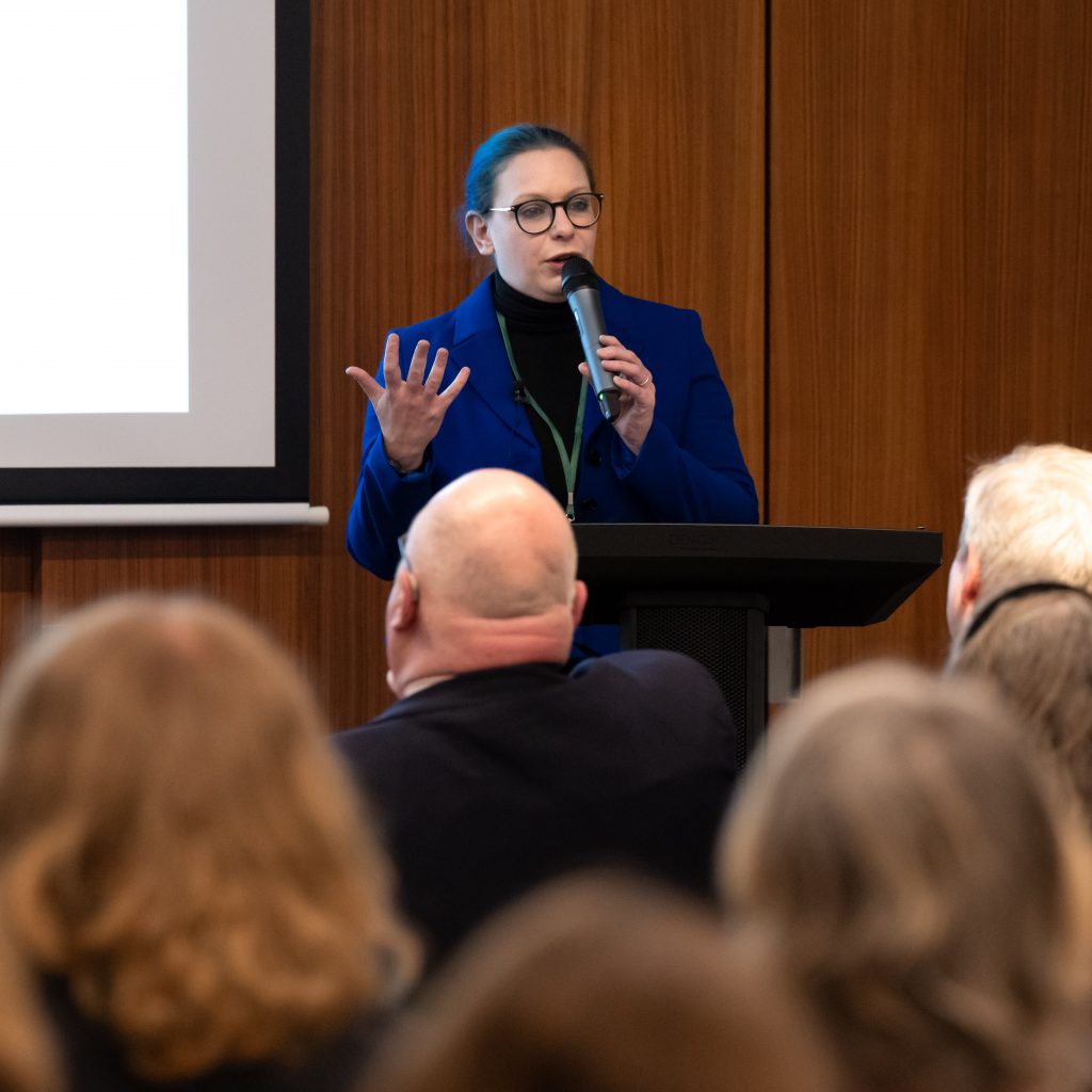 Speaker in a blue jacket presenting at a podium in front of an audience.