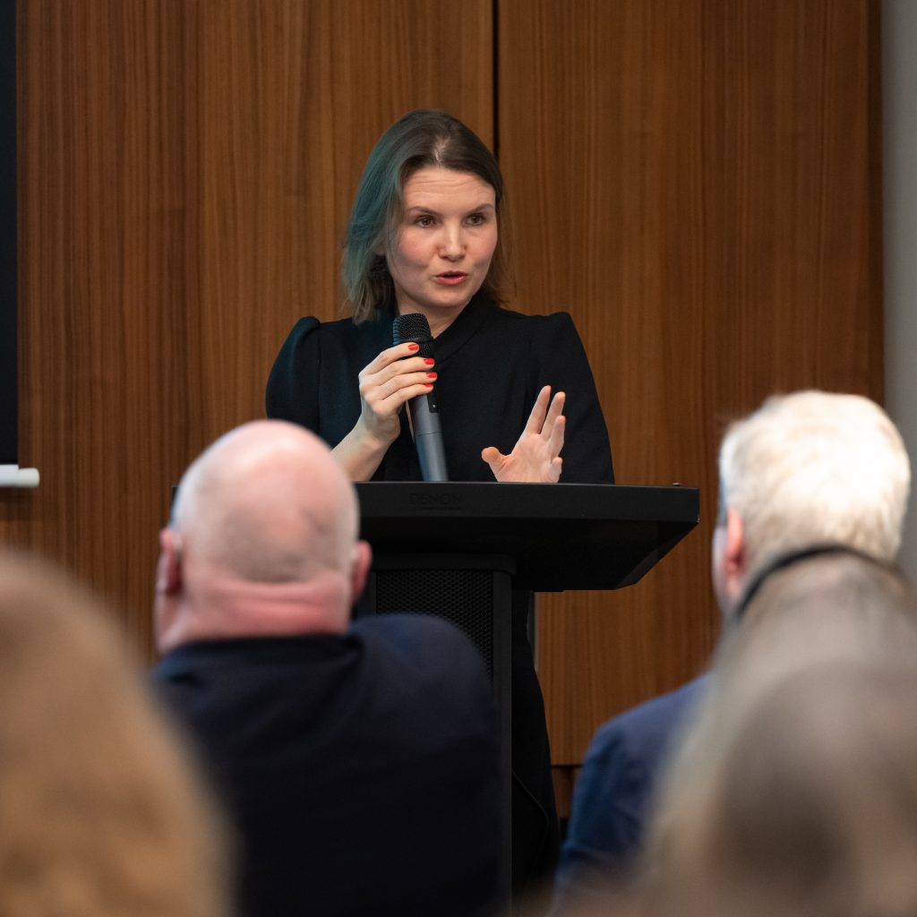 Speaker in a black outfit presenting at a podium, gesturing with one hand.