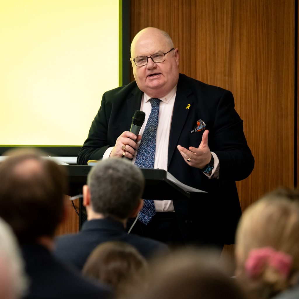 Speaker in a dark suit and tie speaking at a podium with a yellow background.